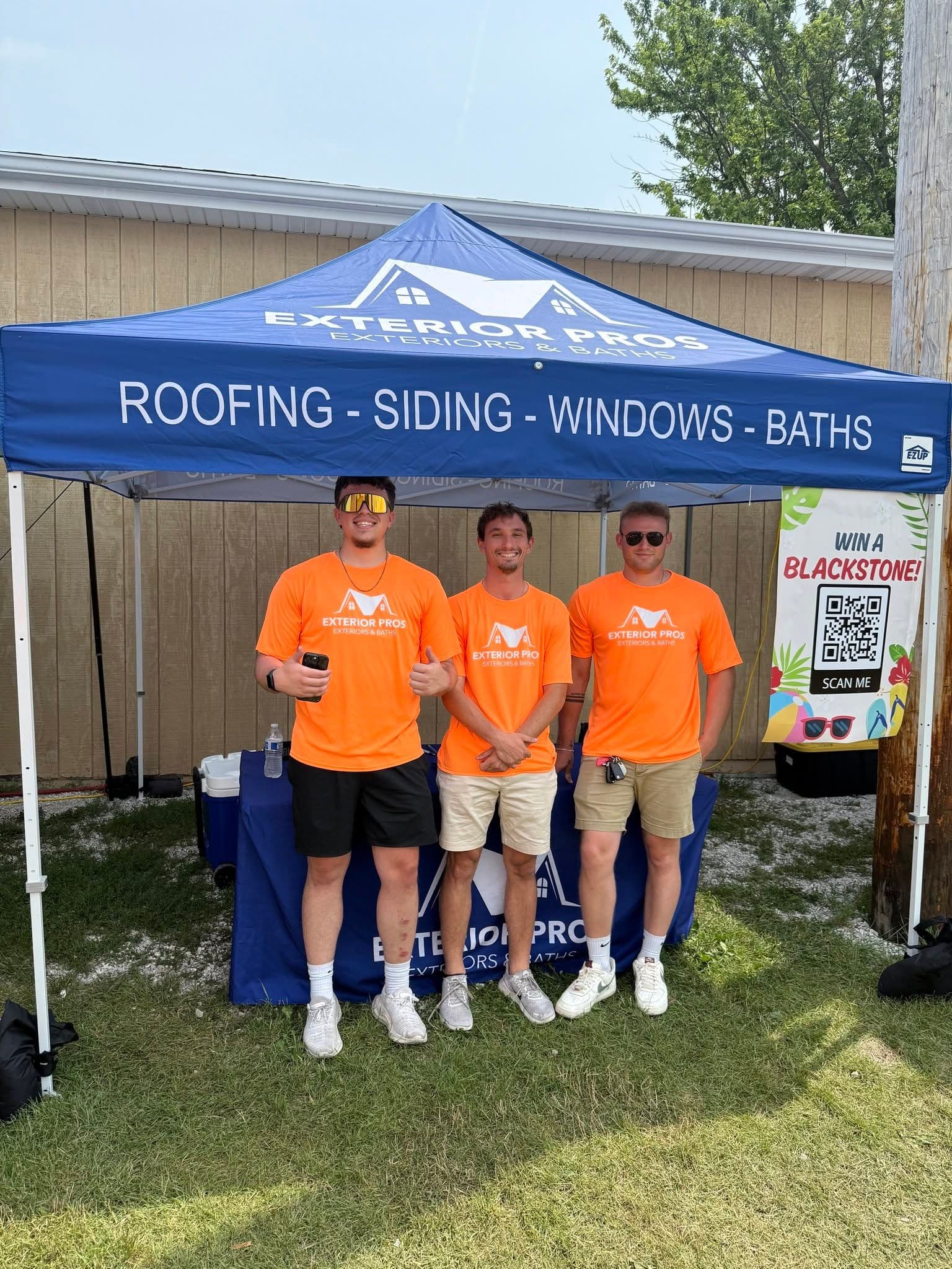 Three Exterior Pros team members in orange shirts standing under a blue promotional tent at an outdoor event
