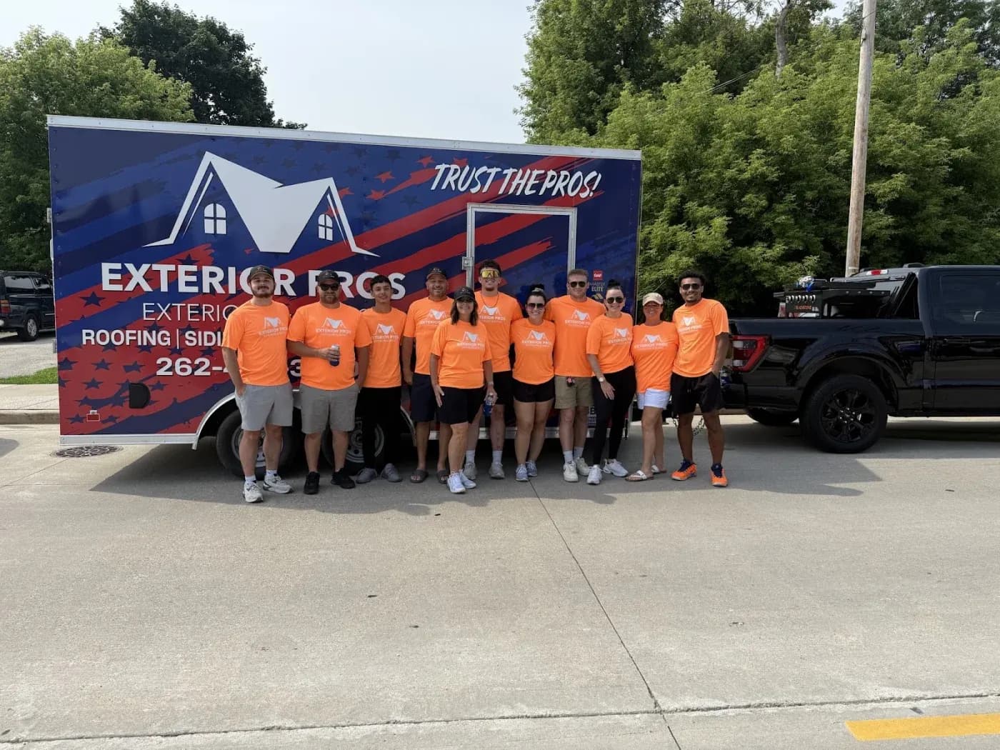 Team of workers in orange shirts posing in front of Exterior Pros roofing company truck and trailer