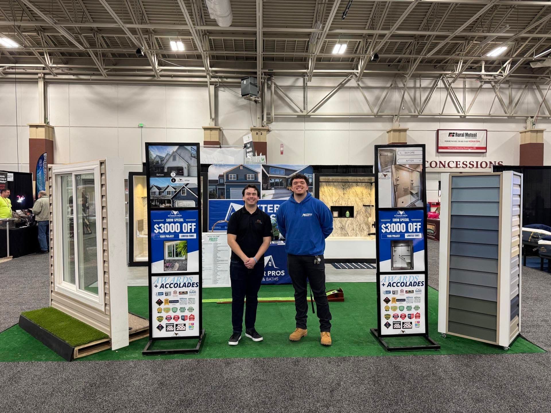 Two staff members standing at a home improvement trade show booth displaying window and door products with $3000 off promotions and award badges