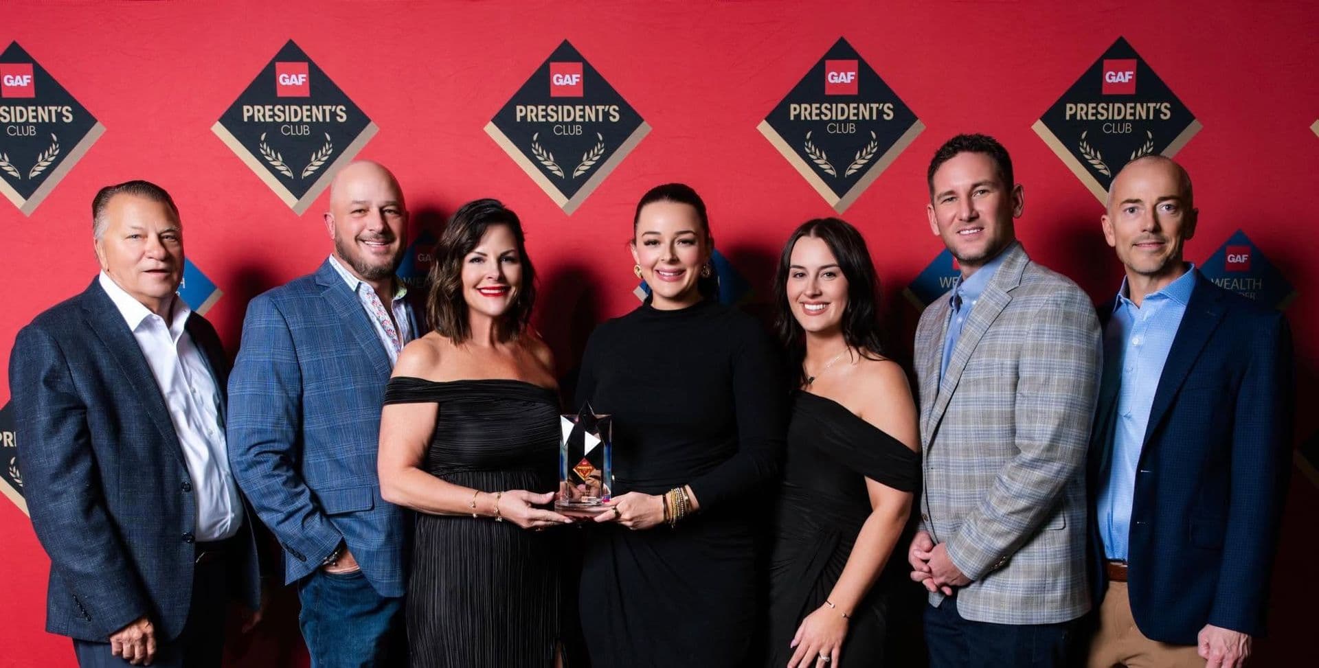 Seven people posing with an award at a GAF President's Club event against a red backdrop with repeating diamond-shaped logos