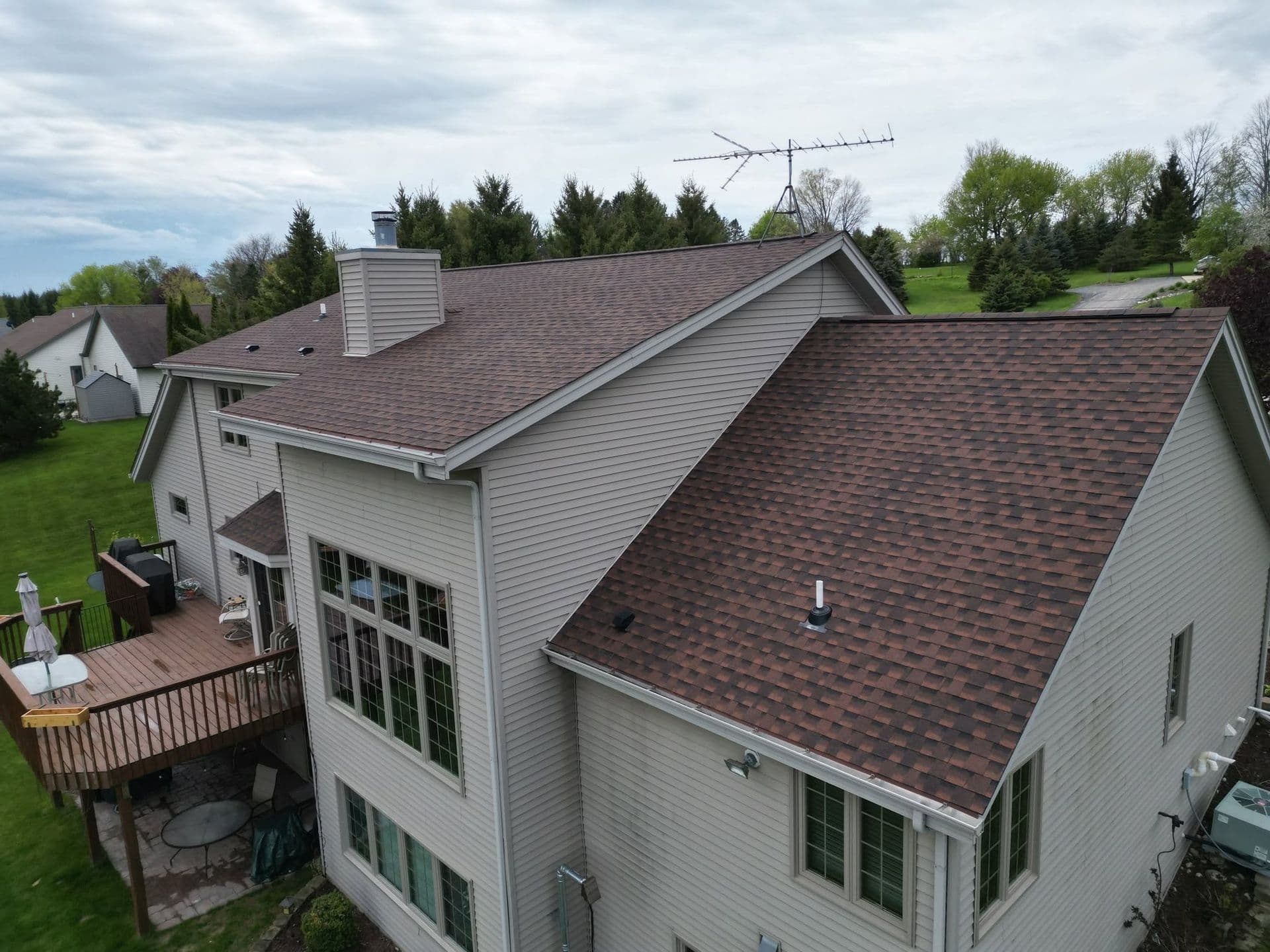 Aerial view of a two-story residential home with brown shingle roof, white siding, deck, and TV antenna, surrounded by green lawn and evergreen trees