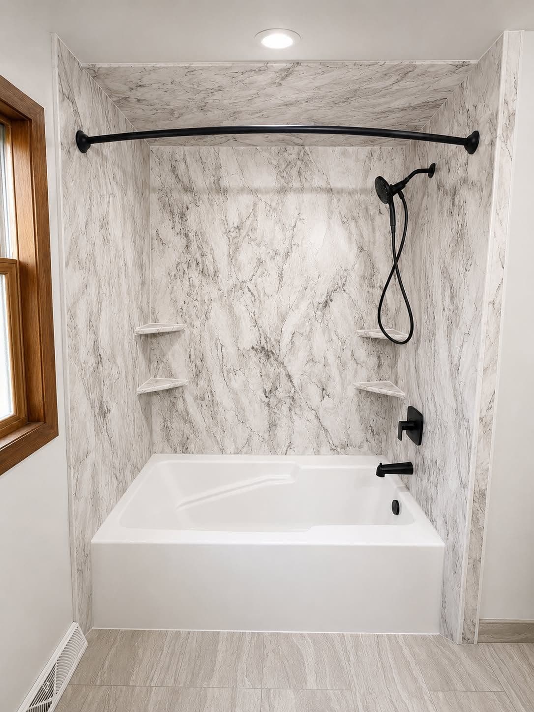 Modern bathroom shower alcove with white marble-look walls, white soaking tub, curved black curtain rod, handheld showerhead, and recessed shelving