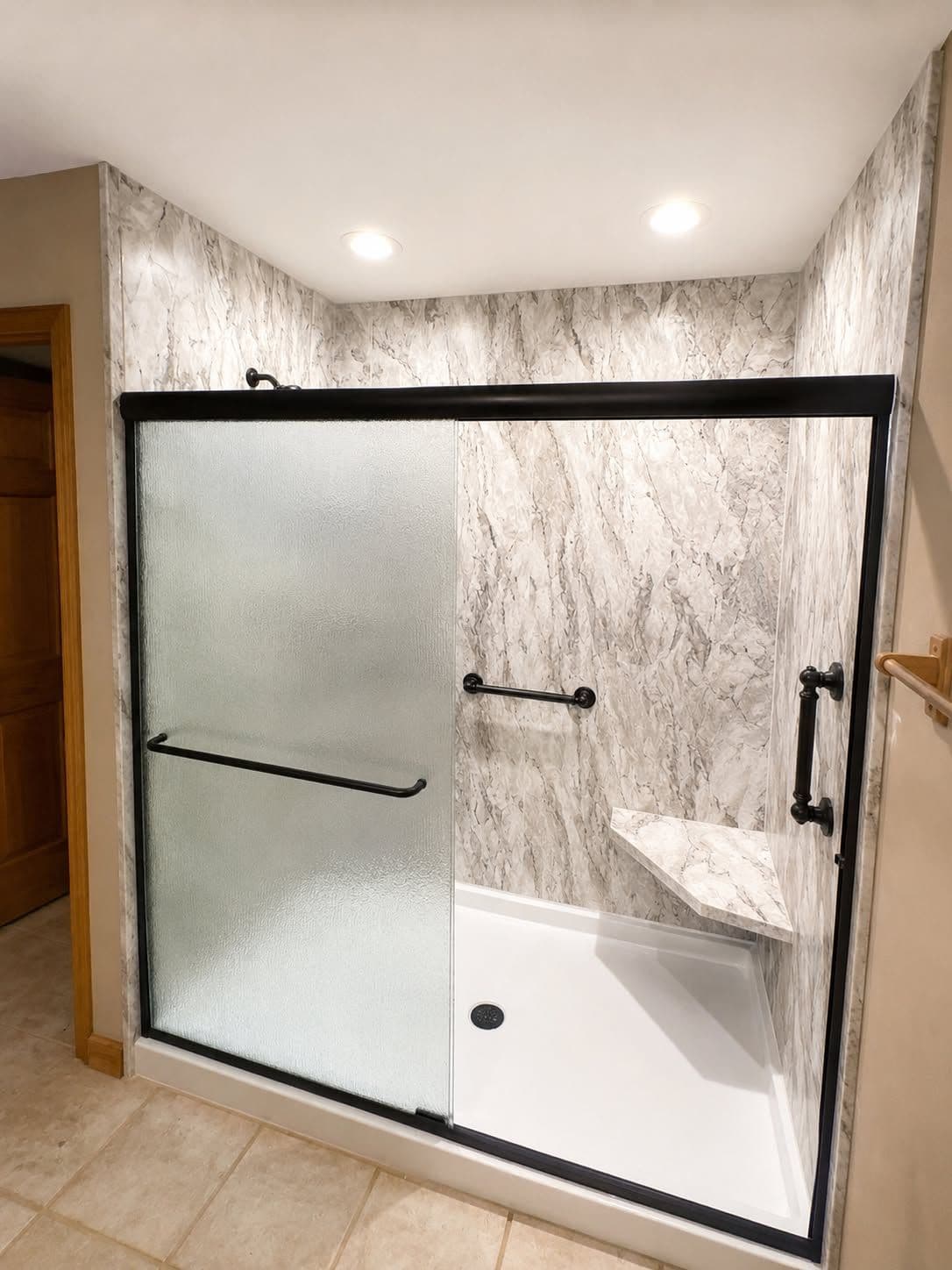Frameless glass shower enclosure with black metal trim and handles, featuring marble-tiled walls and a white base pan with built-in shelf