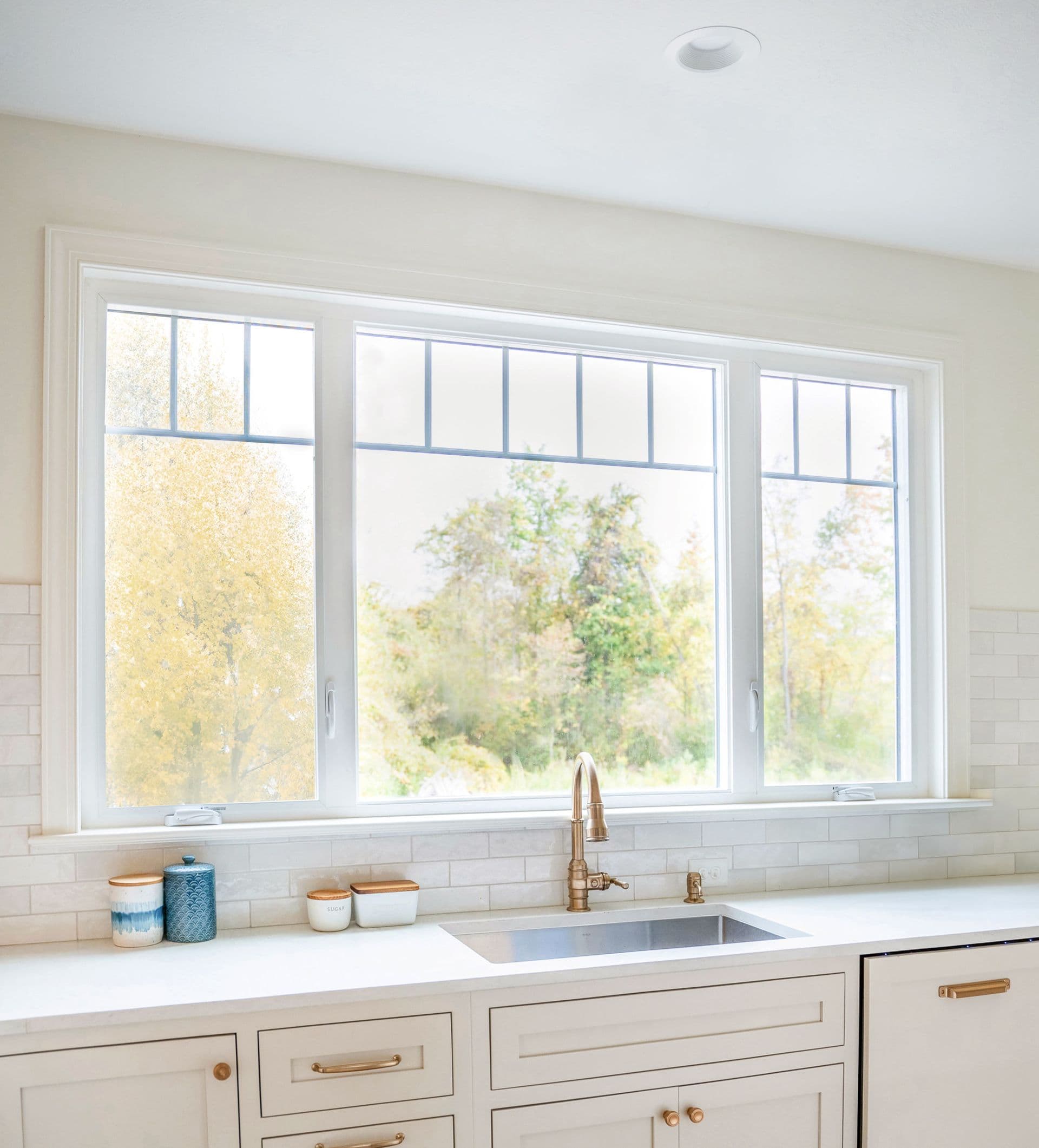 Kitchen sink with brass faucet under three casement windows overlooking autumn foliage and trees - Provia