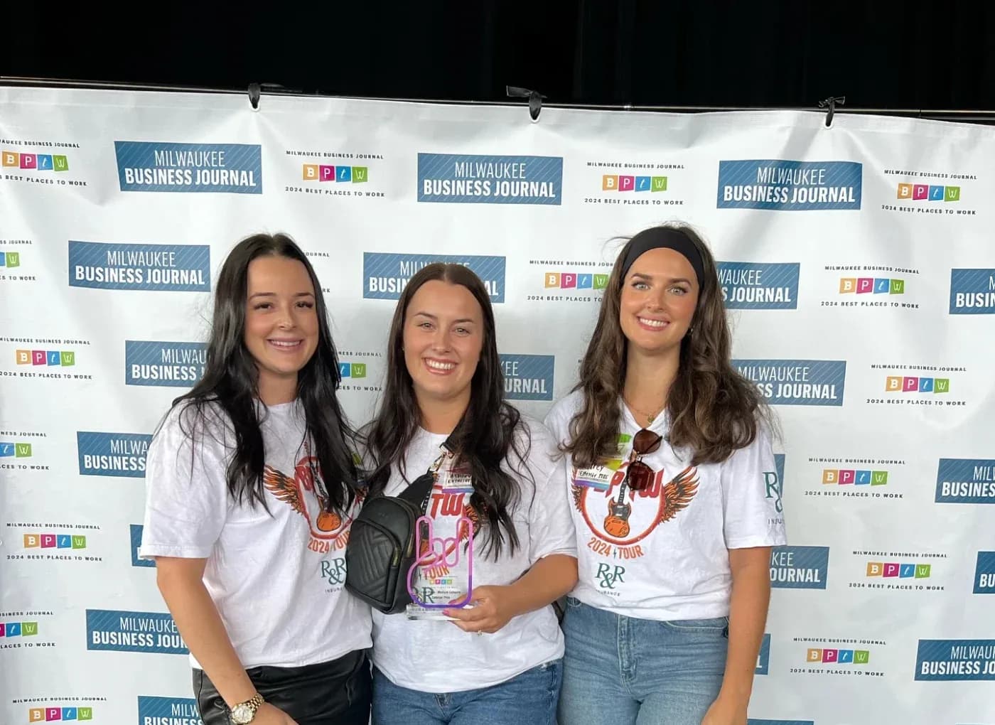 Group photograph of three women smiling in front of a white backdrop branded with "Milwaukee Business Journal 2024 Best Places to Work" logos. They are wearing matching white graphic T-shirts, and the woman in the center is holding an award trophy shaped like a hand giving a rock-on sign.