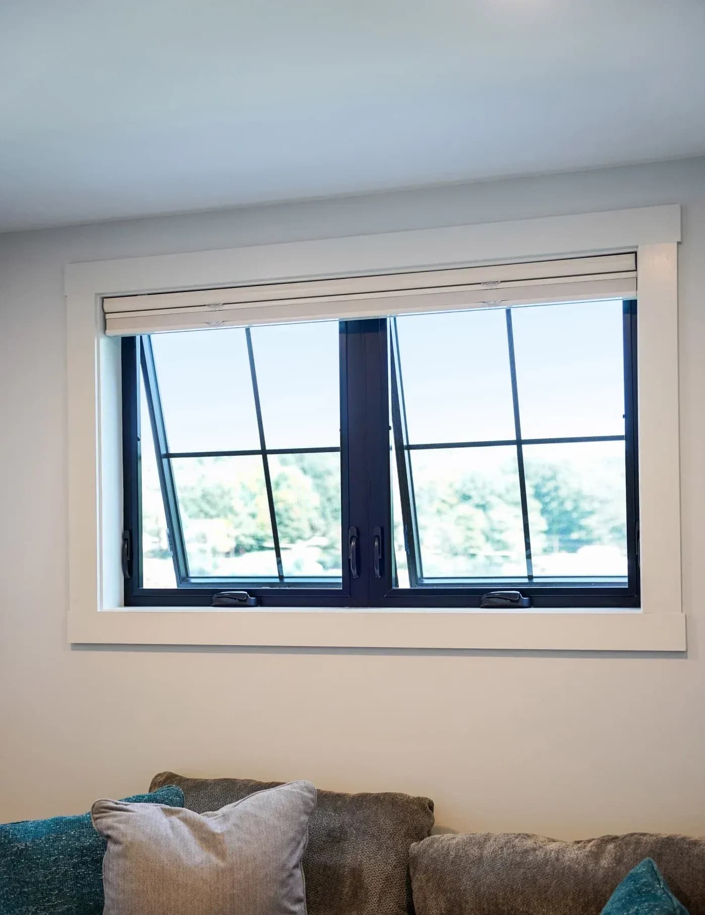 Interior view of a two-lite awning window set featuring dark black frames with simple grid lines, installed with clean white wooden trim casing on a light grey wall above a plush sofa. The dual window sashes are cranked open slightly at the bottom, swinging outward.