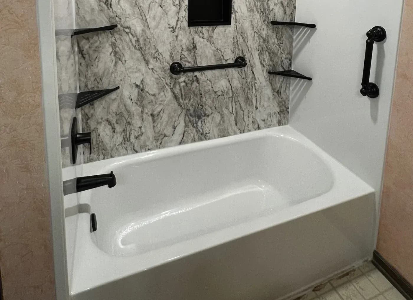 Modern bathroom with white soaking tub, gray marble tile surround, black metal fixtures, and floating shelves above the tub