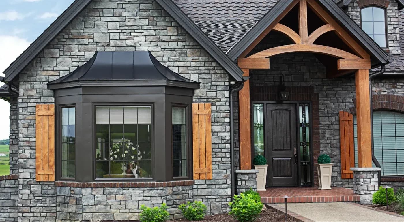 Modern stone house exterior with bay window, wooden shutters, arched entryway with exposed timber beams, and planters flanking the dark wooden door