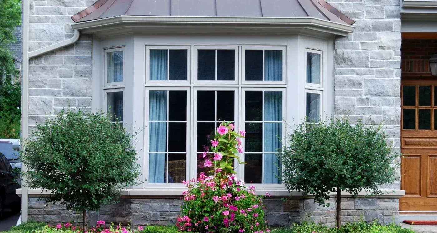 Stone house exterior with large multi-pane window, pink flowering shrubs in front garden, and trimmed green bushes on either side of the window