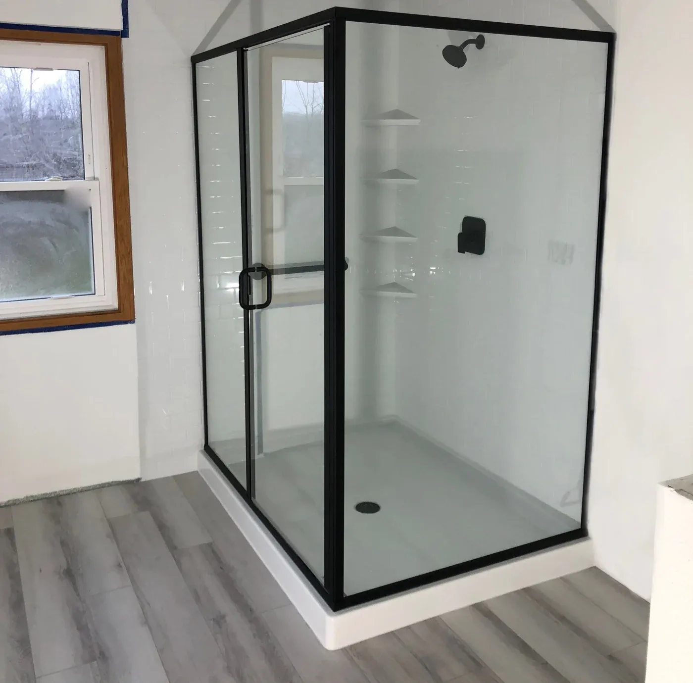 Modern bathroom with a glass-enclosed shower featuring black trim. There are light wood floors, a window, and white walls, conveying a clean, minimalist feel.