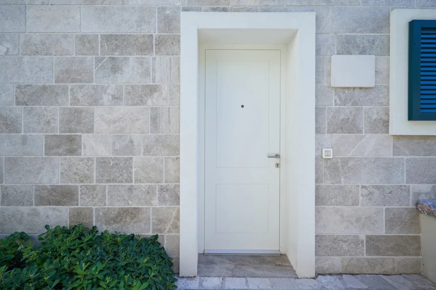 Frontal exterior view of a modern, minimalist white entrance door with a silver handle set within a thick white casing. The door is set against a light grey stone block wall, featuring green leafy shrubs on the left and a glimpse of dark blue window shutters on the upper right.