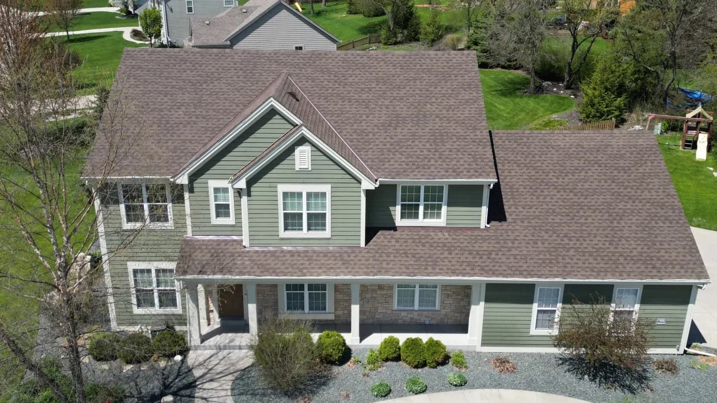 Aerial view of a two-story residential home with gray-brown asphalt shingle roof, green and stone siding, white-trimmed windows and columns, front porch, and landscaped yard with shrubs and mature trees