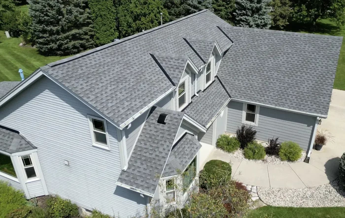 Aerial view of residential home with gray asphalt shingle roof, white siding, two-car garage, and manicured landscaping including shrubs and lawn