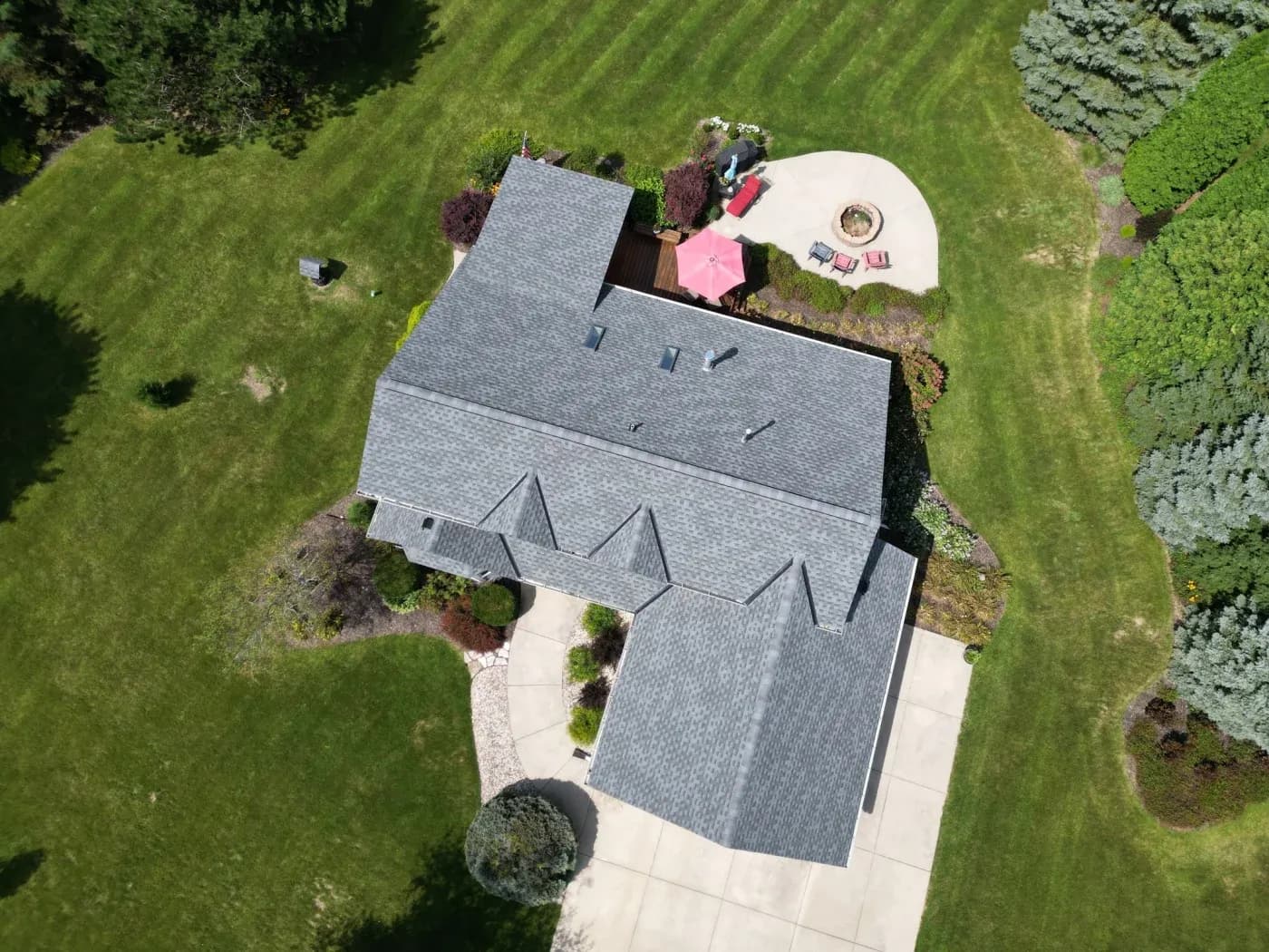 Aerial view of residential home with gray shingle roof, curved patio with pink umbrella and seating, surrounded by manicured lawn and evergreen landscaping