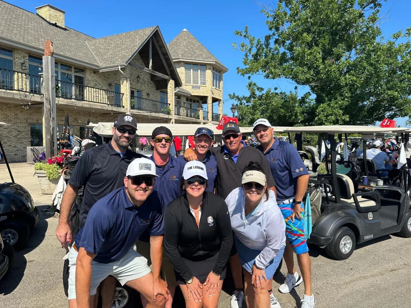 Group of eight golfers posing together at a charity golf event, with golf carts and a brick mansion in the background