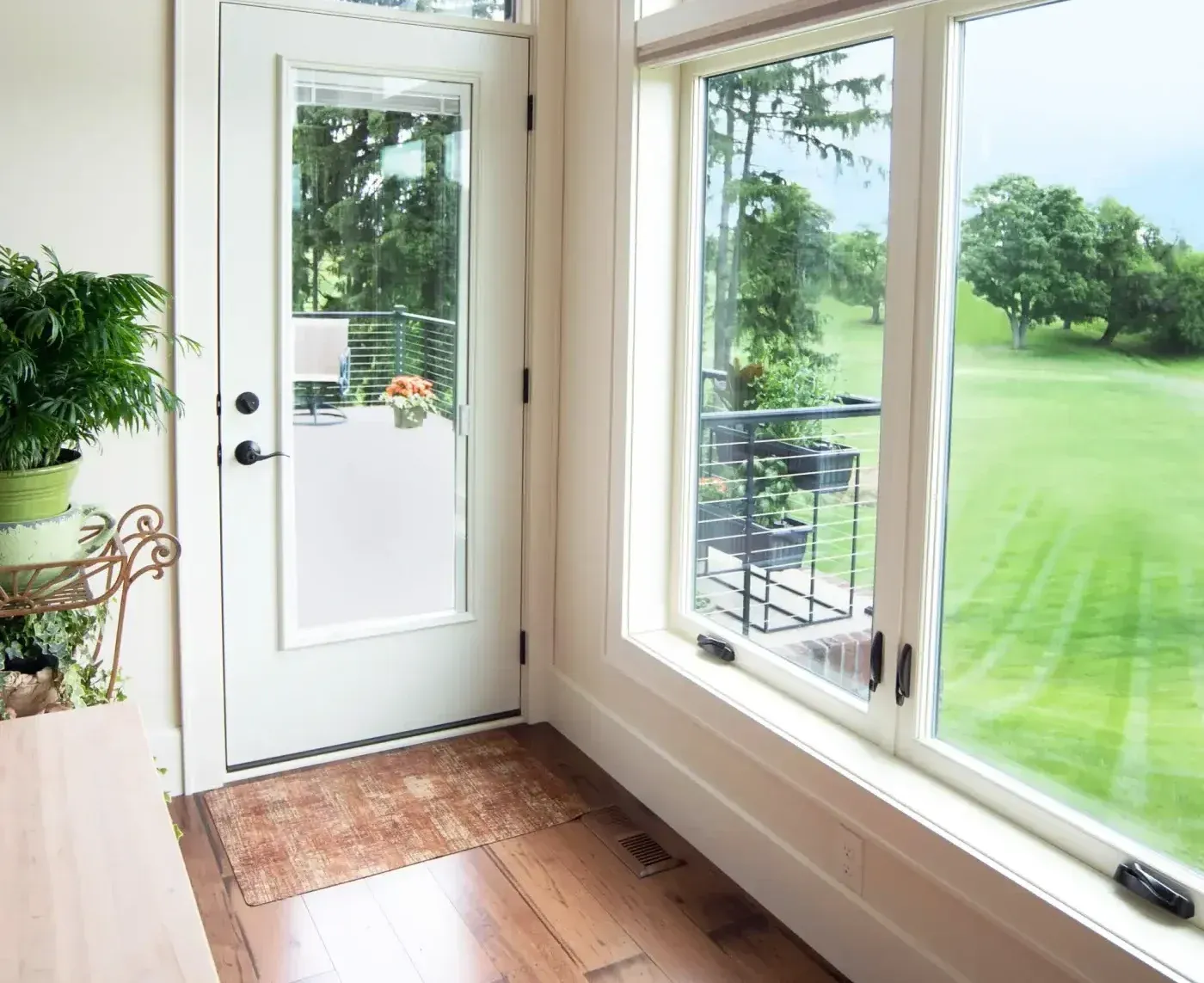 Bright mudroom with white door and large windows overlooking green lawn, wooden floor, potted plants on shelves, and metal railing on deck outside