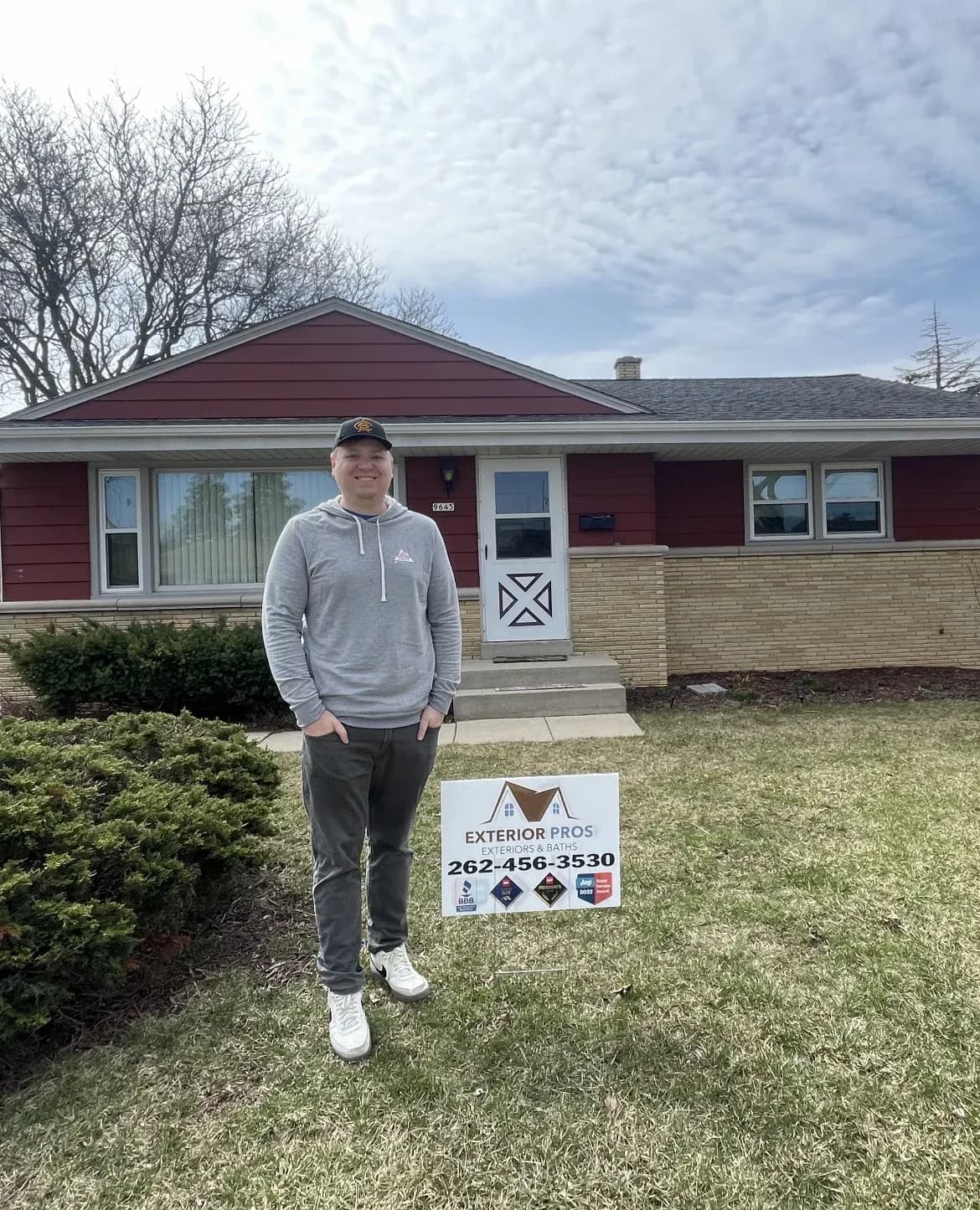 Man standing in front of a single-story red and tan house holding an Exterior Pros yard sign with contact information
