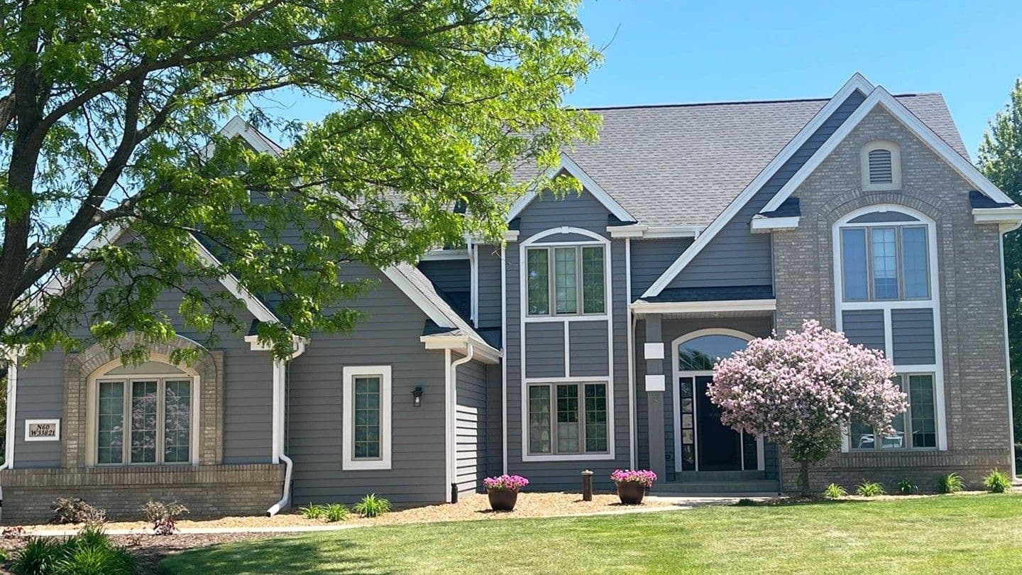 Two-story residential home with brick and siding exterior, multiple gabled rooflines, large windows, flowering magnolia tree, landscaped front lawn, and mature shade tree on left side
