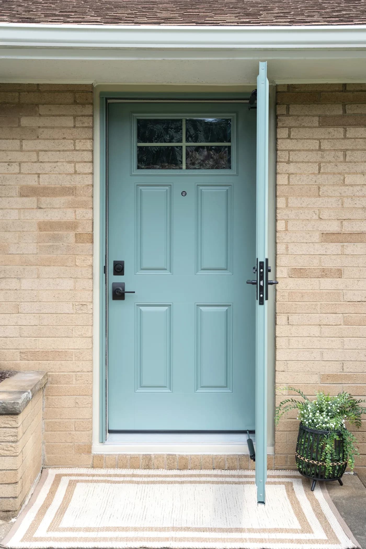 Light blue front door with six panels and window transom, dark hardware, set in brick entryway with striped doormat and potted plant