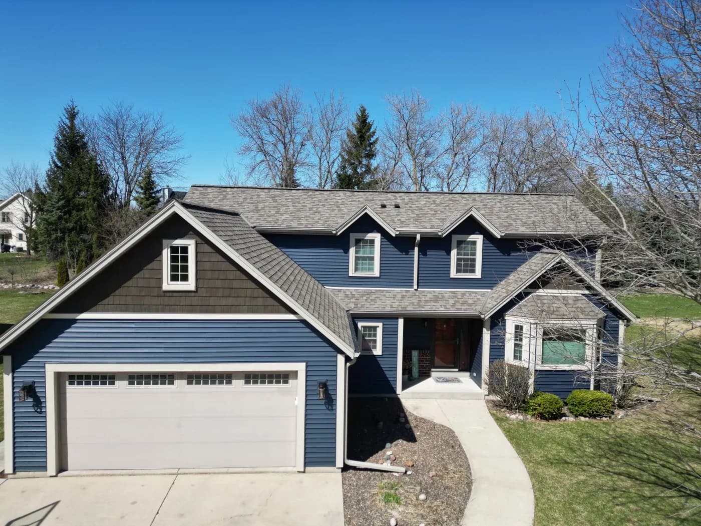 Two-story blue house with white trim, two-car garage, and multiple gabled roof sections, surrounded by bare trees and green lawn