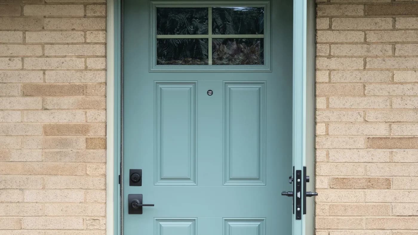Teal front door with four raised panels and transom window, flanked by tan brick walls, featuring black door handle and deadbolt lock