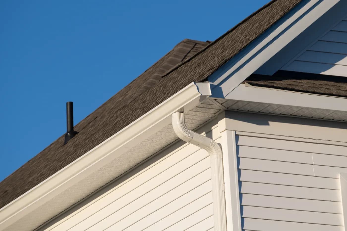 Close-up of house roof corner showing dark wood shingles, white fascia trim, gutter downspout, and clear blue sky
