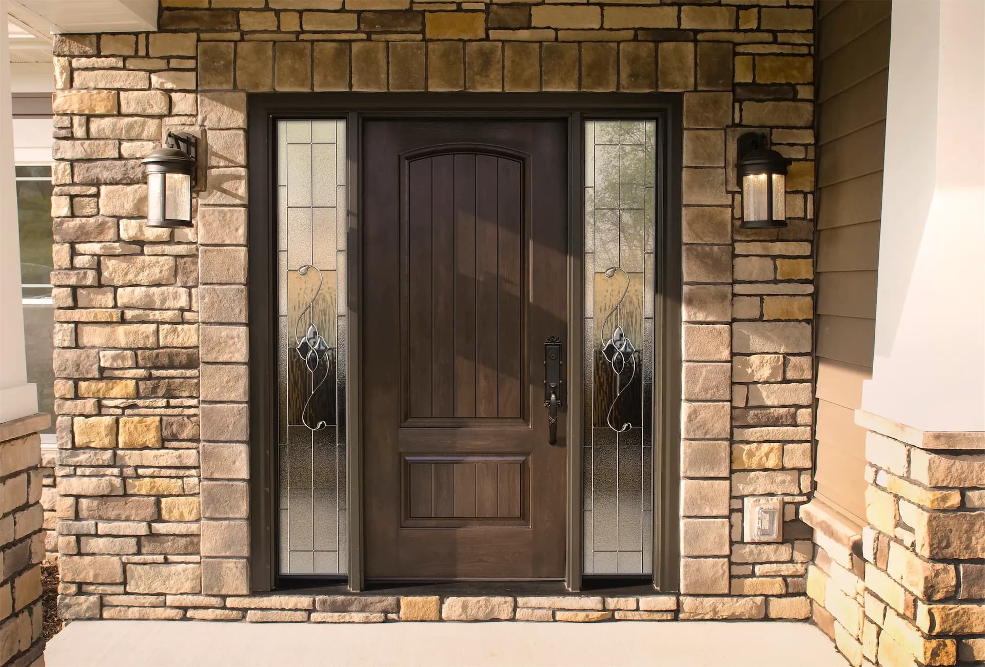 Exterior view of a dark wood front entrance door with decorative leaded glass sidelights, flanked by wall sconces and set into a stacked stone wall facade.