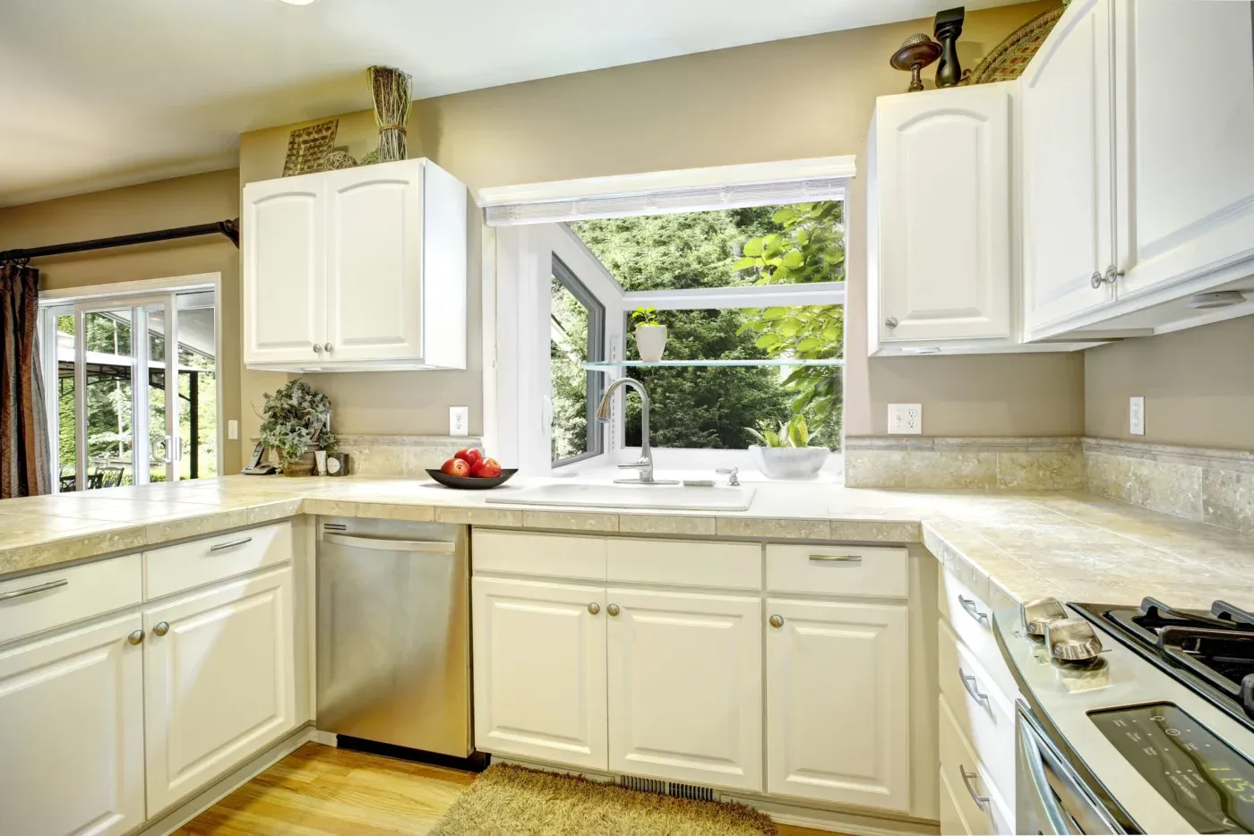 Interior view of a kitchen featuring white upper and lower cabinetry, beige tile countertops, stainless steel appliances, and a vinyl garden window installed above the sink with a glass shelf holding small plants overlooking lush green trees.