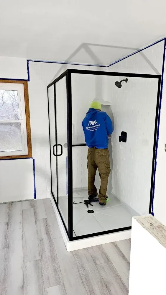 Person installing a black-framed glass shower enclosure in a bathroom with white tile walls and gray wood-look flooring