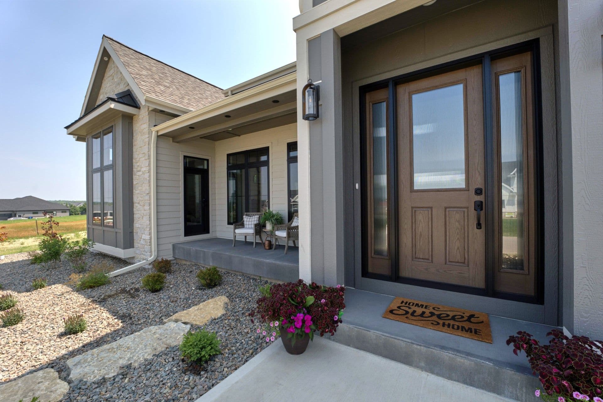 Modern home exterior with covered front porch, wood entry door with sidelights, stone and siding facade, landscape with gravel and flowering plants, and home sweet home doormat