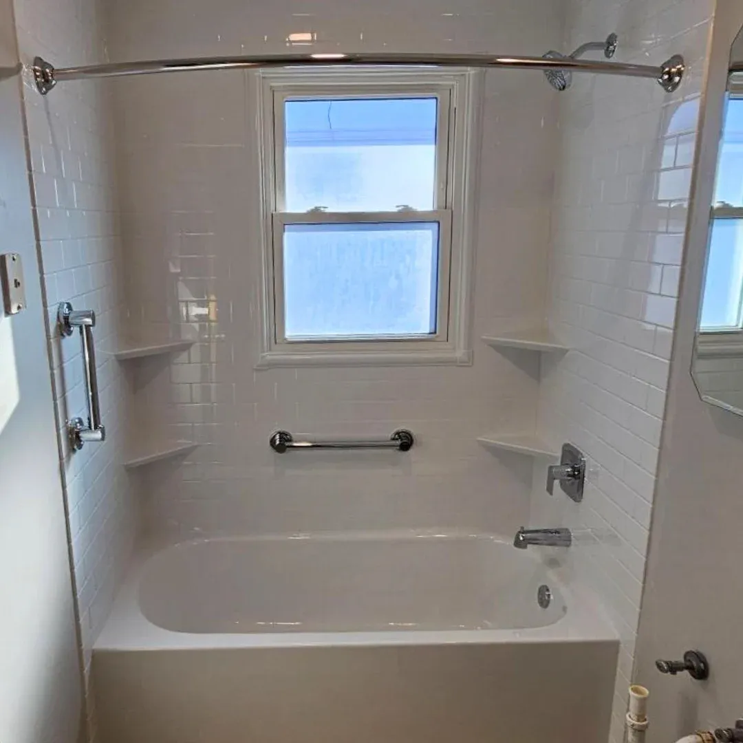 Bathroom shower enclosure with white subway tile walls, curved chrome curtain rod, built-in shelves, and a window with blue sky visible outside