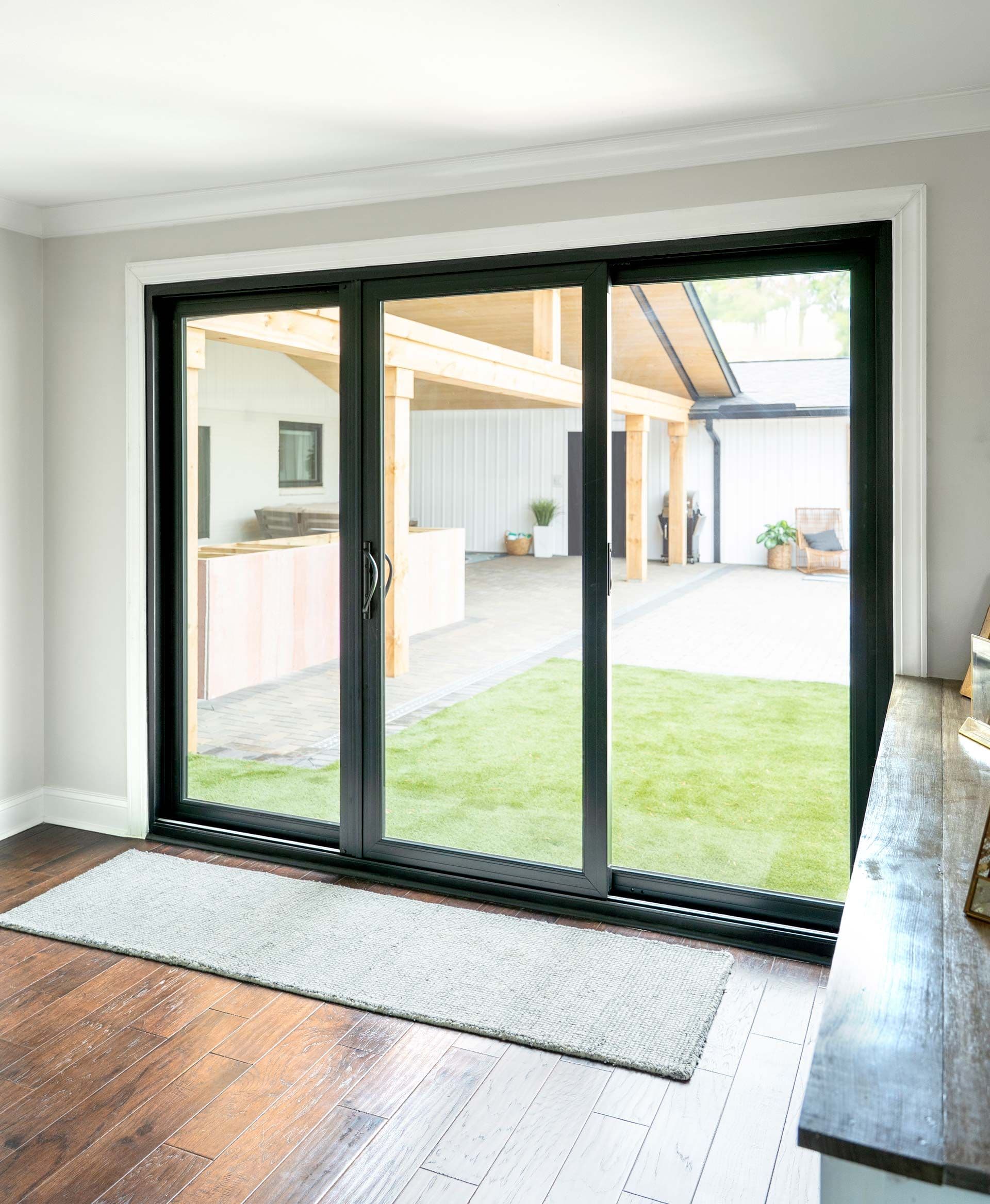 Three-panel black framed sliding glass doors opening from a wood and tile floored room to a backyard patio and garden