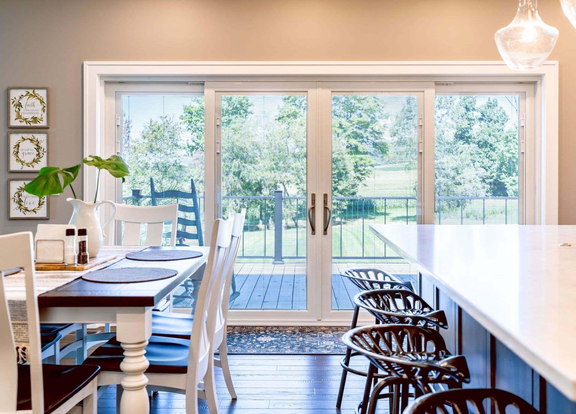 Bright dining area with white chairs at a dark table, multi-panel glass doors opening to a deck overlooking trees, inspirational wall art, and kitchen counter with bar seating