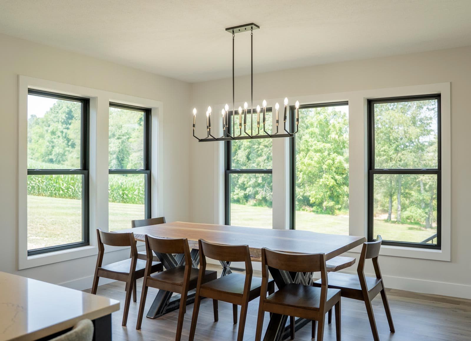 Modern dining room with rectangular table, six wooden chairs, and linear black chandelier suspended from ceiling above table, flanked by four large windows overlooking green landscape - provia