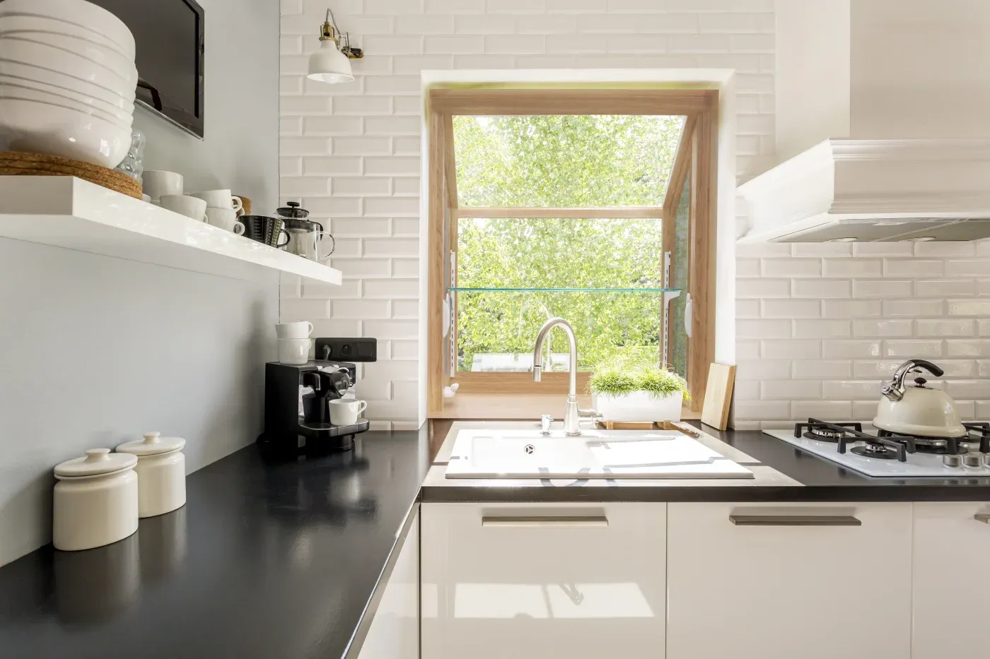 Modern kitchen with white subway tile walls, white cabinetry, black countertops, sink with faucet, coffee maker, stovetop, and large window overlooking green foliage
