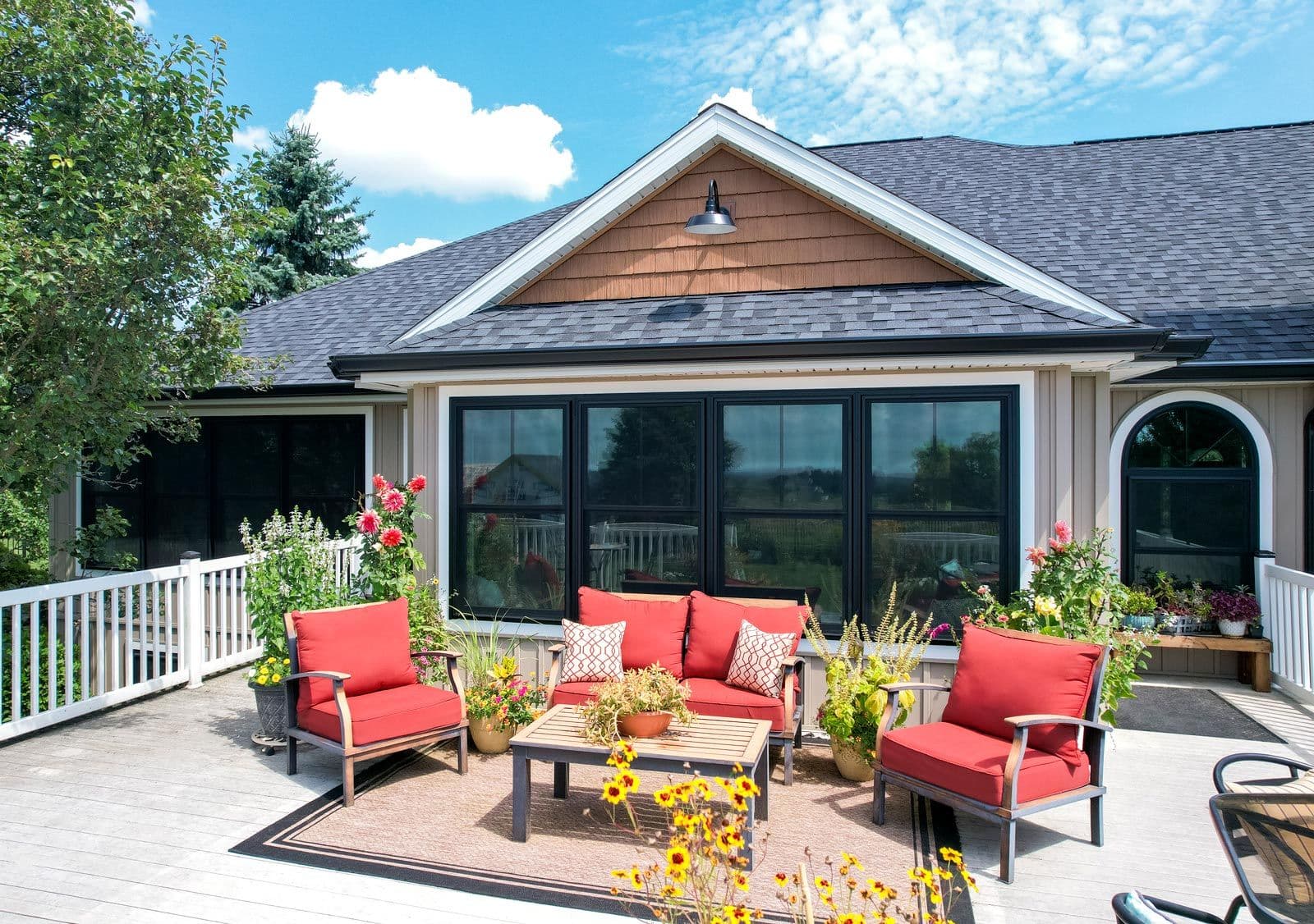 Modern home exterior with patio seating area featuring red cushioned chairs and sofa, potted flowers, and large black-framed sliding glass doors opening to the deck