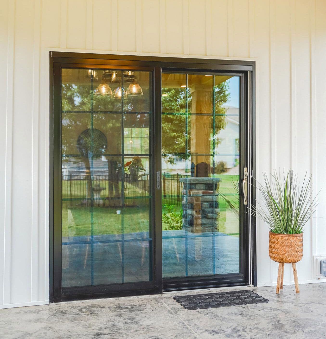 Black aluminum sliding glass patio doors opening to a waterfront lawn and garden, with pendant lights visible inside and a potted plant beside the doors