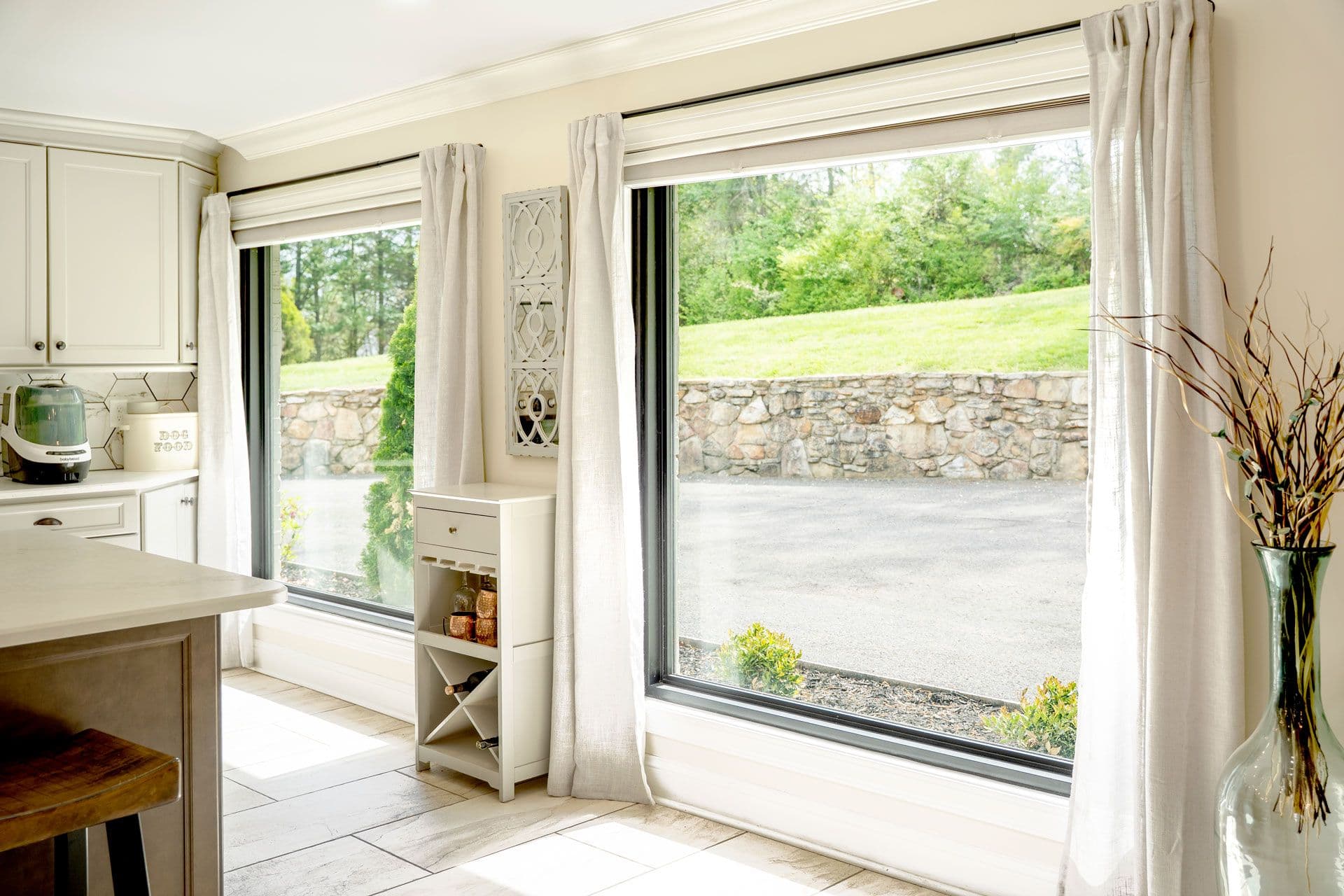 Picture Window - Modern kitchen with cream cabinetry and island opening to outdoor patio through large sliding glass doors, overlooking stone wall and green lawn with forest beyond - Provia