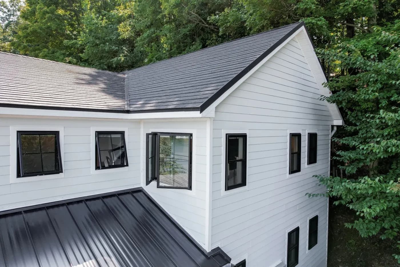 Modern two-story white house with dark metal roof and black trim windows, surrounded by dense green trees