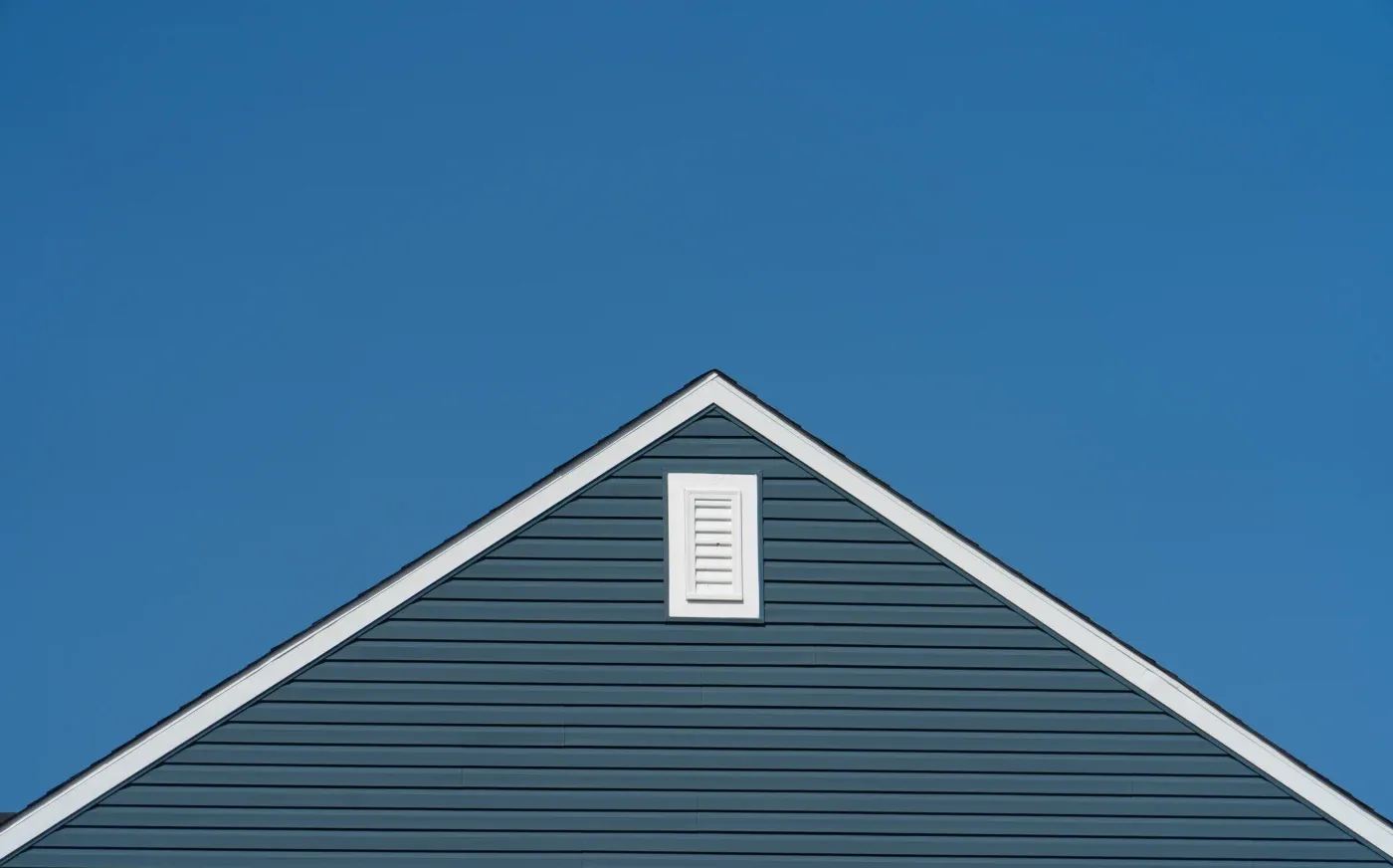 Symmetrical exterior view of a triangular roof peak with dark slate blue horizontal lap siding, crisp white trim along the rake board, and a small rectangular white louvered attic vent set against a clear blue sky.