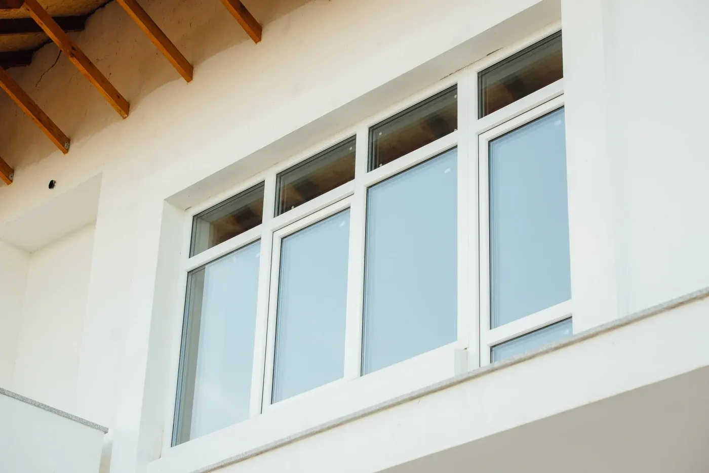 Low-angle exterior view of a multi-pane white-framed window installed into a smooth white stucco building wall, featuring exposed wooden roof rafters visible along the upper left ceiling trim.