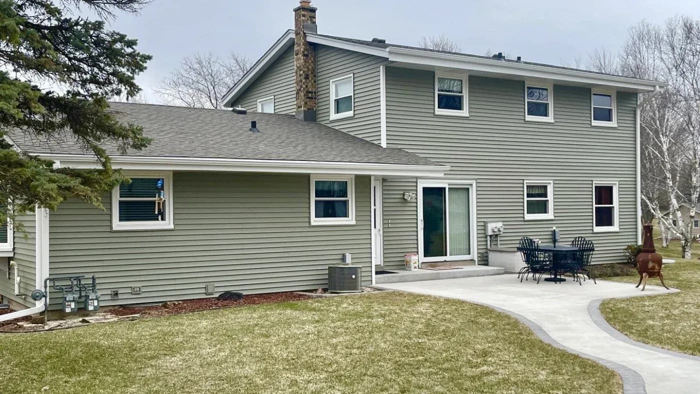 Two-story green and gray siding residential house with stone chimney, curved concrete patio with Adirondack chairs, manicured lawn, and bare trees in spring setting