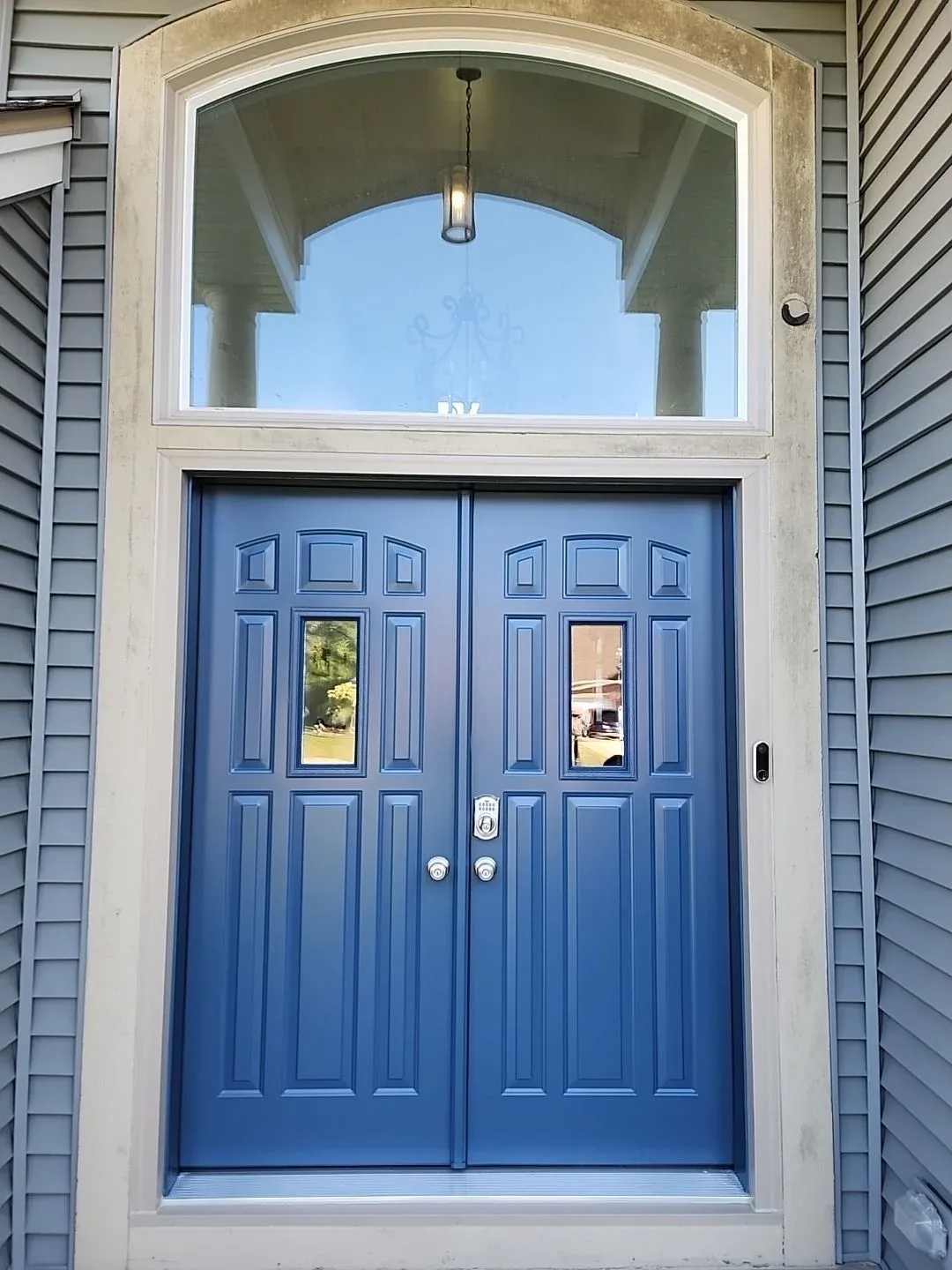 Blue double doors with glass panels and transom window above, flanked by gray siding on home entryway