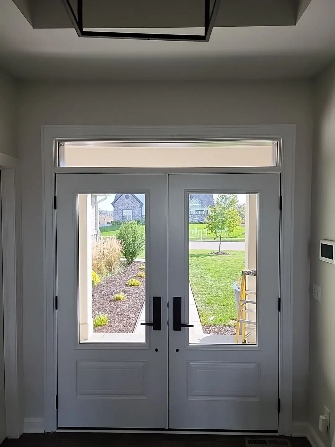 Double glass doors with transom window opening to suburban backyard with landscaping, lawn, and neighboring houses
