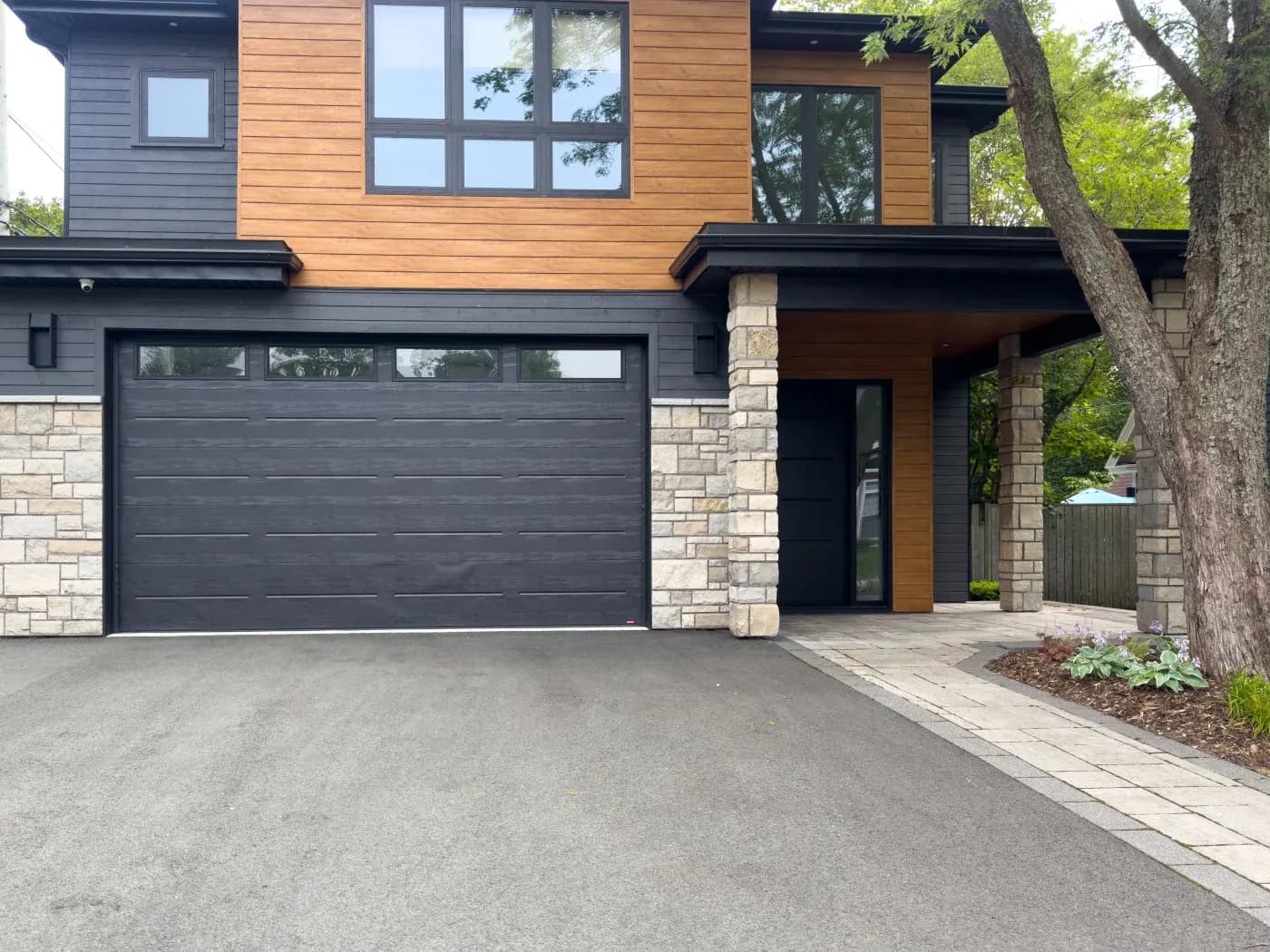Modern two-story home with dark garage door, stone and wood siding, large windows, and curved driveway lined with pavers