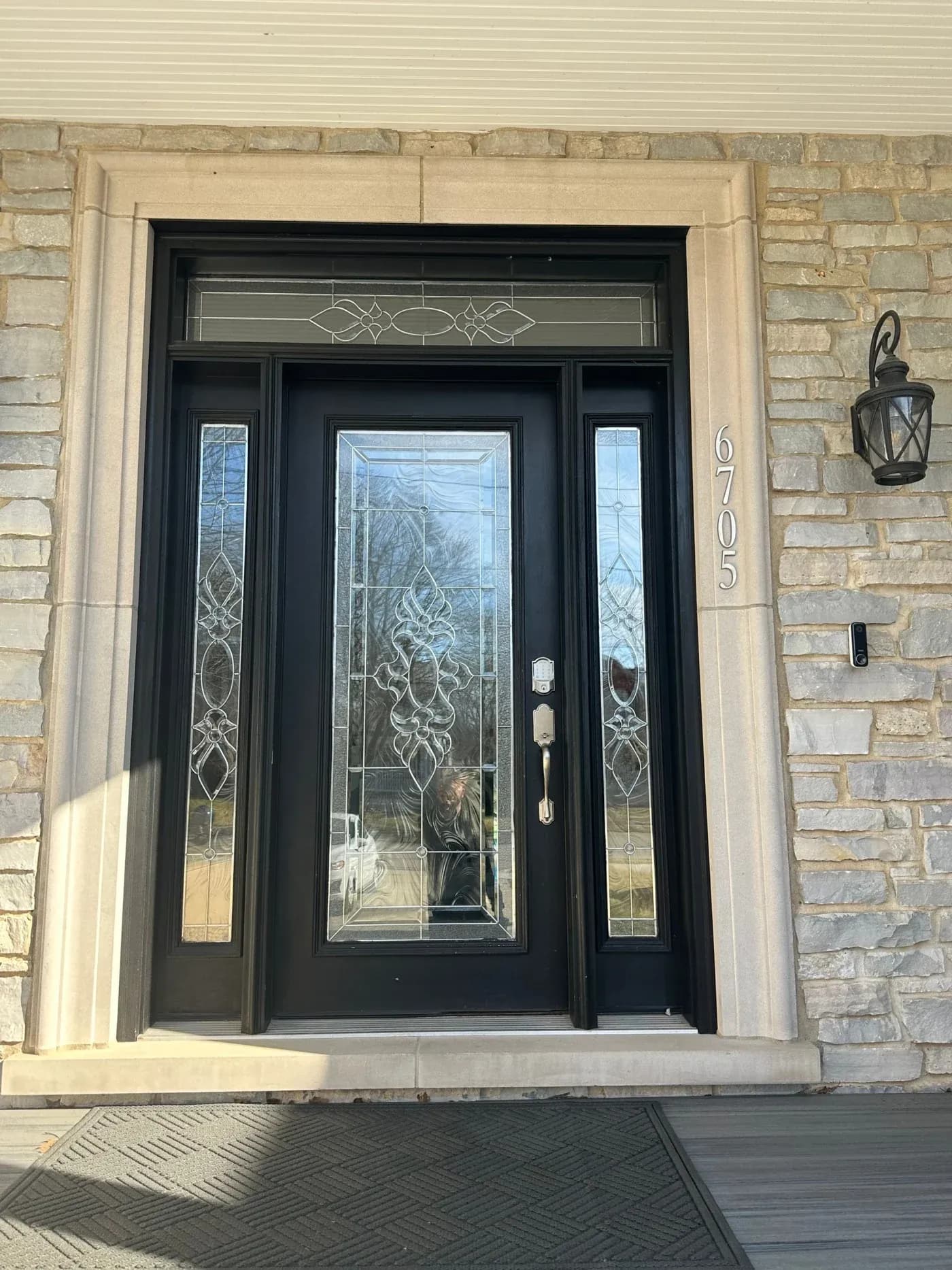 Black front door with decorative glass panels and sidelights, brass house number 6705, and lantern sconce on tan brick exterior