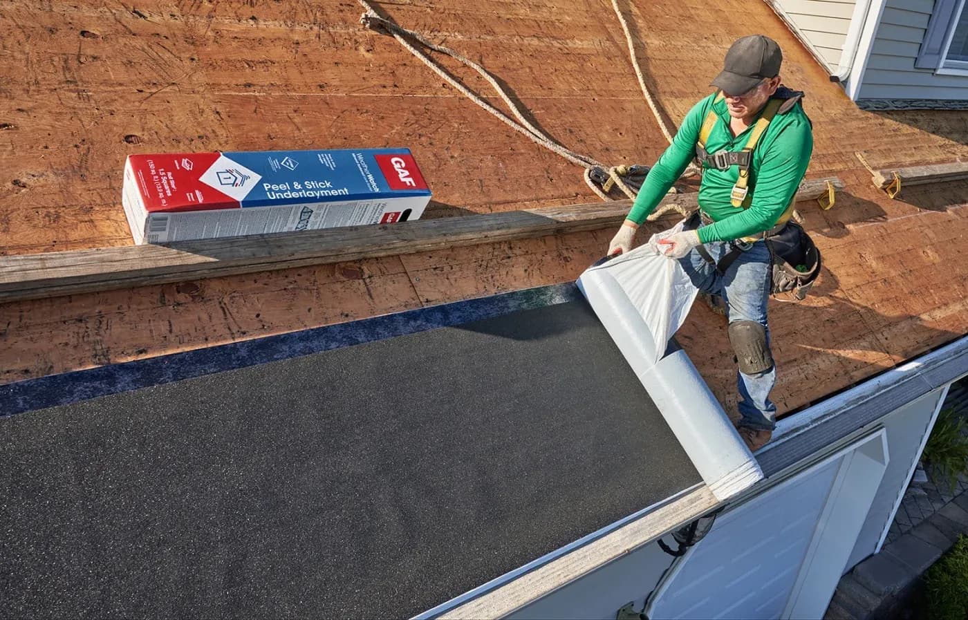 Roofer in green shirt applying peel and stick underlayment to roof sheathing during residential roofing installation