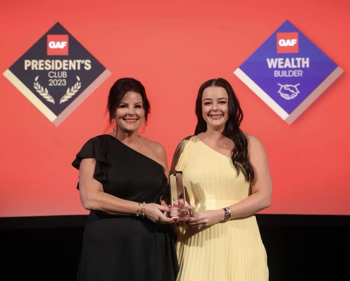 Two women holding an award on stage in front of QAF President's Club 2023 and Wealth Builder badges on a coral background