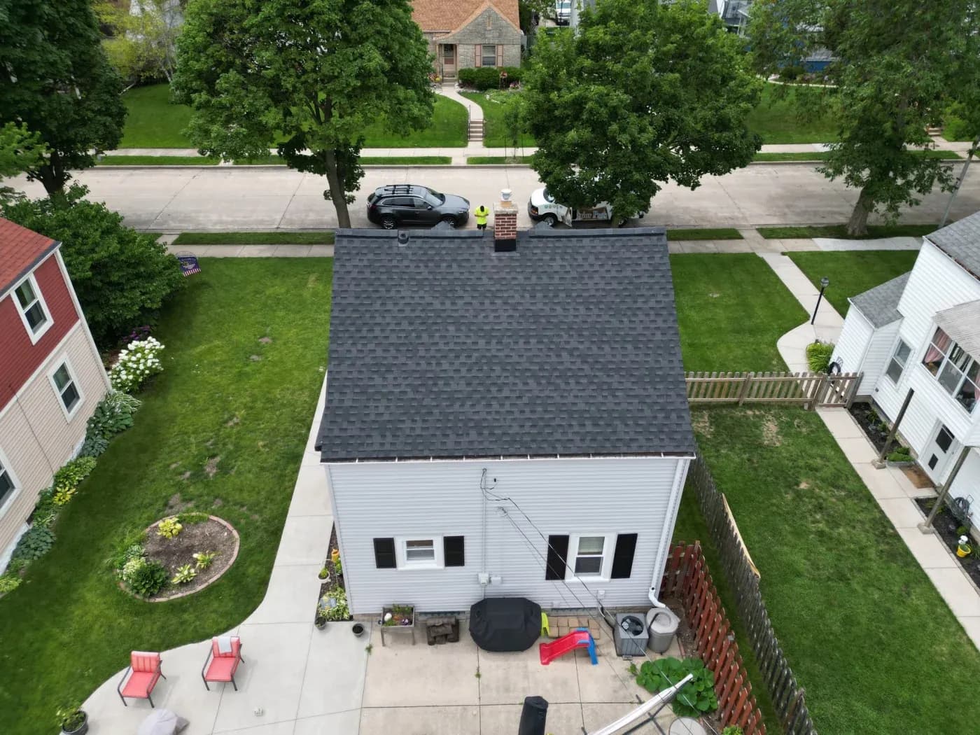 Aerial view of a single-story house with gray shingled roof and white siding, surrounded by neighboring homes and mature trees on a residential street