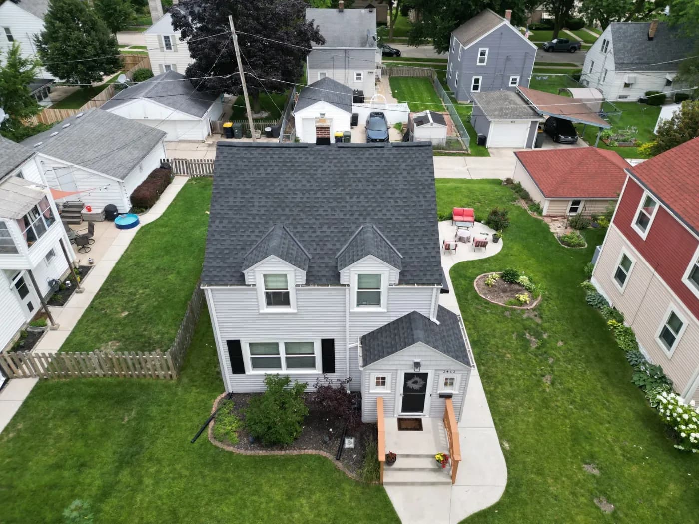 Aerial front view of a two-story residential home with light grey horizontal siding and a dark charcoal GAF Timberline HDZ asphalt shingle roof. The house features two dormer windows on the second floor, black shutters on the ground floor window, a small front entrance porch with steps, a paved walkway, and a green yard set in a suburban neighborhood.
